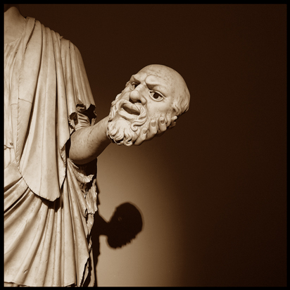 Photo by Augusto De Luca, of detail of statue. Apparent is a torso draped in a toga, with an arm and, in the hand, there is a face, perhaps a mask.