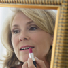 Blond adult woman, shown in a mirror, putting on lipstick.