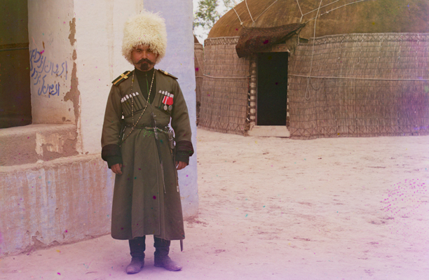 Soldier in tall white fur hat and dressed in old fashioned dark green uniform, standing in front of a house and a yurt.