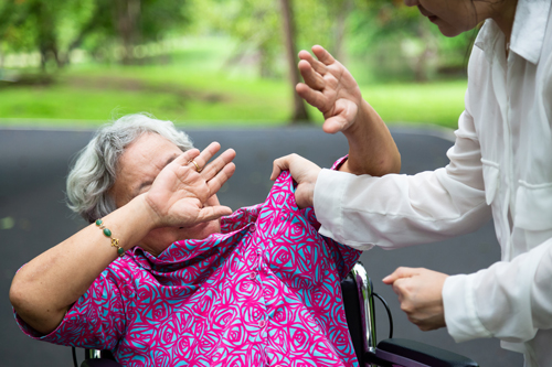 Woman with white hair and pink smock in a wheelchair, attendant in white coat grabbing her smock with another hand in a fist illustrating article by Richard Klass about nursing homes